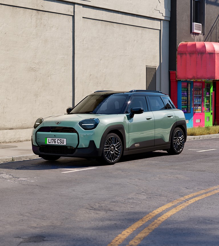 Gray MINI Aceman parked on a city street beside a bright purple and red building, featuring modern design and black roof.